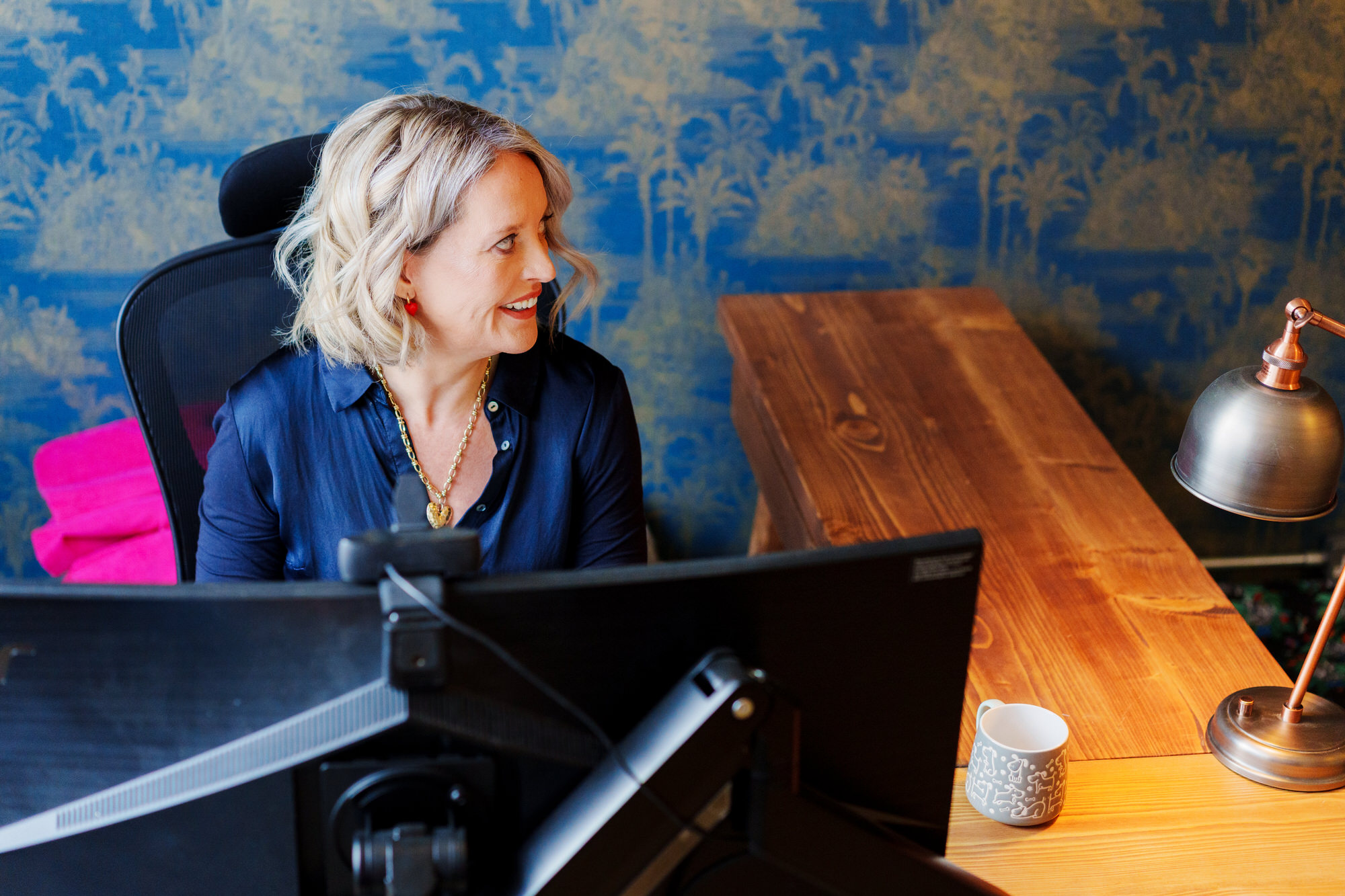 Sarah Morgan-Kelly at her desk in the Lewes office of Morgan Kelly Solicitors, in front of the firm's deep-navy palm-leaf wallpaper.